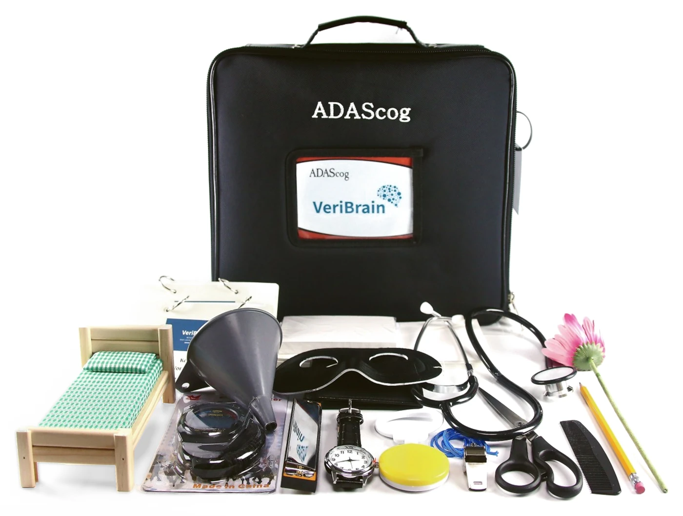 VeriBrain 라벨이 붙은 검은 ADAScog 가방과 그 앞에 놓인 검사 도구들. 나무 침대 모형, 깔때기, 눈가리개, 청진기, 손목시계, 가위, 호루라기, 꽃, 연필, 빗이 보인다